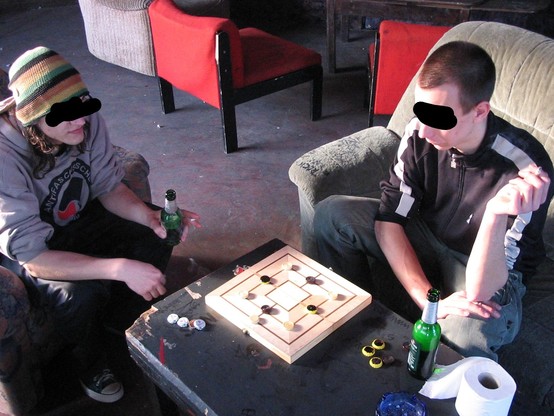 Zwei junge Männer spielen Mühle. Sie trinken dabei Bier und rauchen. Die Gesichter wurden mit schwarzen Balken unkenntlich gemacht. 