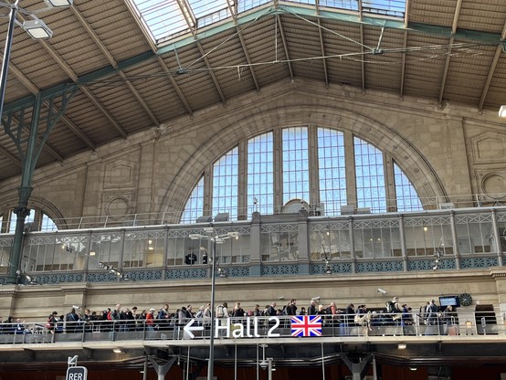 A queue of people above a sign saying Hall 2 and a union flag. Large windows of Gare du Nord behind 