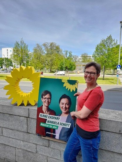 Ulrike Caspary präsentiert ein Plakat mit den Portraits von Manuela Schott und sich selbst, daneben das Symbol der Sonnenblume.