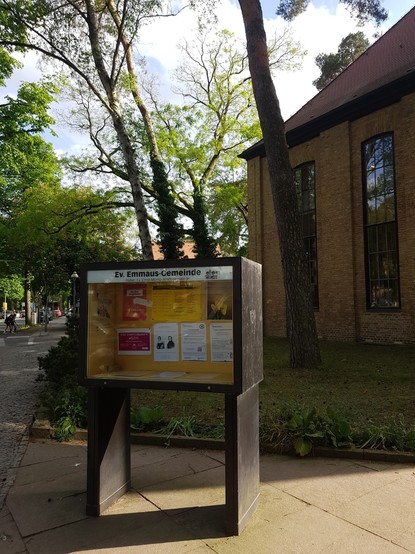 Schaukasten der Gemeinde, vor gelben Hintergrund hängen Hinweise, im Hintergrund Bäume und der untere Teil eines Kirchen Gebäude