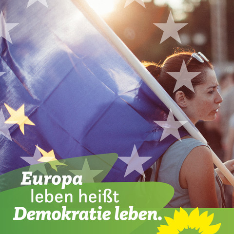 Auf dem Bild zu sehen ist eine Frau im Seitenprofil. Auf ihrer Schulter hat sie eine große Europaflagge. Unten links steht auf grünen Blättern: Europa leben heißt Demokratie leben.