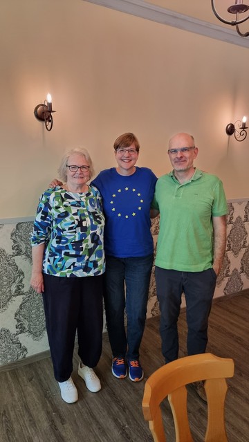 Three people smiling for the camera in a room with wall sconces and patterned wallpaper. Michaela Reimann in the middle is wearing a EU t-Shirt