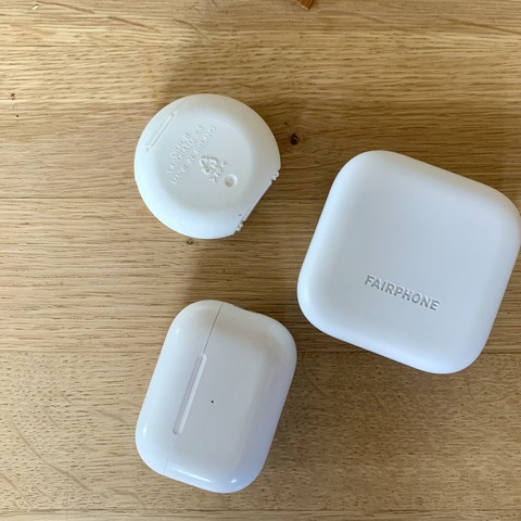 Drei weiße Döschen auf einem Holztisch. Zwei davon sind eckige Ladeboxen für drahtlose Ohrstöpsel. Eins davon ist Rund und eigentlich Zahnseide.
