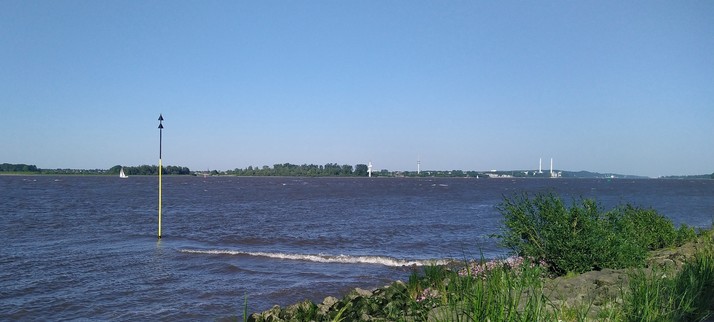 Blick über die Elbe in Richtung Hamburg. Das Wasser hat Schaumkronen auf den Wellen. Ein Segelboot ist zu sehen. Auf dem gegenüberliegenden Ufer sind die zwei Schornsteine in Wedel sichtbar.