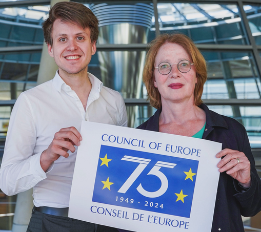 Der grüne Bundestagsabgeordnete Max Lucks und seine Kollegin Tabea Rößner halten zusammen ein Plakat hoch, auf dem eine große 75 auf blauem Untergrund mit gelben Sternen steht. Darüber steht Council of Europe. 