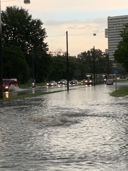 A flooded street with cars driving through the water, a tram in the distance, and an overcast sky.