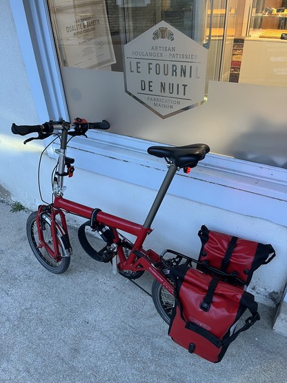 Red Birdy bike with red panniers in front of a bakery with a sign that reads:

• L'ATTESTATION -
QUALITE & BANETTE
MENTION D'EXCELLENCE
M. & MME BADET
ARTISAN
BOULANGER - PATISSIER
LE FOURNIL
DE NUIT
• FABRICATION •
MAISON