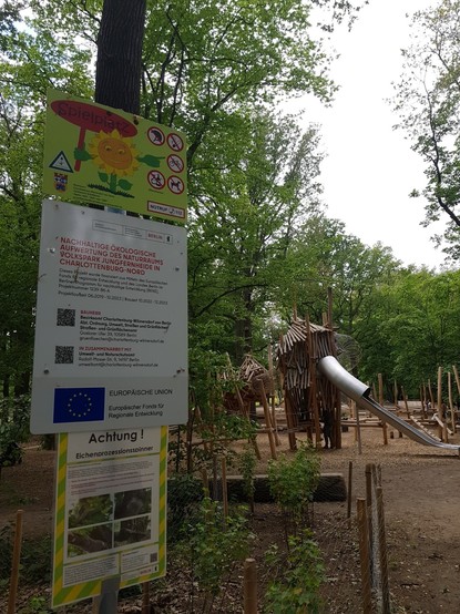 Im Vordergrund steht neben einem bunten Spielplatz Schild ein Hinweis auf EU Förderung mit der Flagge der EU
Im Hintergrund sind viele Spielgeräte aus Holz, eine Rutsche und Bäume mit grünen Blättern zu sehen