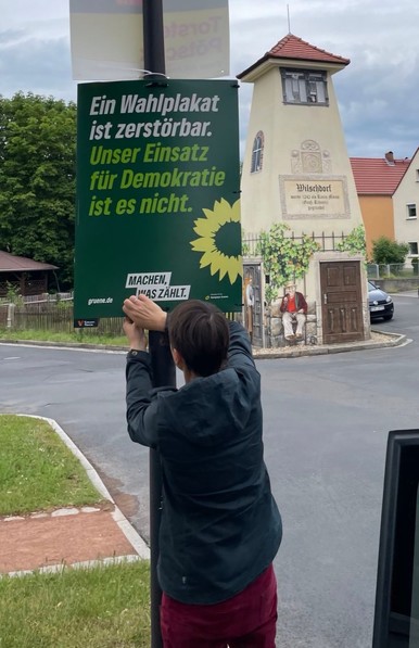 Eine Person hängt auf einem Dorfplatz ein Plakat auf. Text: Ein Wahlplakat ist zerstörbar. Unser Einsatz für Demokratie ist es nicht.