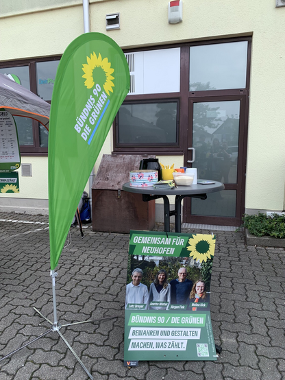 Grüne Beachflag, daneben Stehtisch mit Tassen und Kaffee, davor ein Plakat der Kandidierenden von B90/Die Grünen