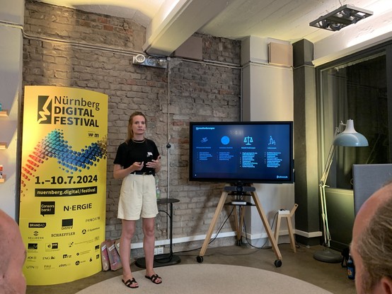 Franziska is presenting at the Nürnberg Digital Festival. In the background, there is a large digital screen displaying information and a yellow banner with festival details for July 2024. The setting has an industrial look with exposed brick walls.