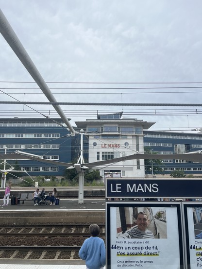 A pic of Le Mans station. There are a series of pylons and cables above the platform that should support canvas but this is missing on the platform nearest the camera. There’s a station sign saying LE MANS and a white building beyond the platforms. 