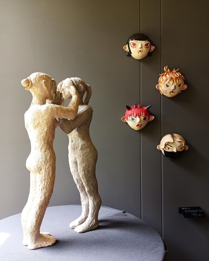 Two tall female ceramic figures touching masks in front of each others faces. In the background there are four colourful ceramic masks hanging on the wall.