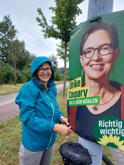 Ulrike Caspary in blauer Regenjacke lächelnd vor einem ihrer Wahlplakate