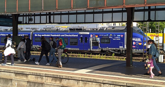 A train platform with people walking along it

Behind is a dark blue train that says it will run between France and Germany on it