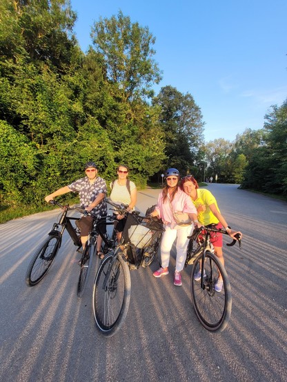 Vier Radfahrende mit ihren Rädern auf einem Kiesweg. Im Hintergrund Wald. Von hinter der Kamera werden Schatten auf die Personen geworfen.