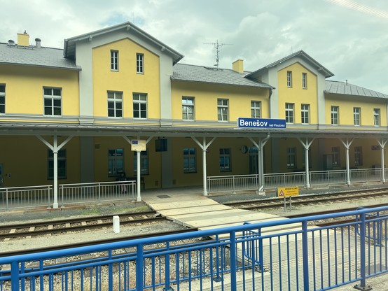 Benešov nad Ploučnicí station building. Yellow painted concrete facade. Good condition. Mix of 2 and 3 storey parts of building. Roof in front of building to provide shelter to passengers. Tracks in front. 