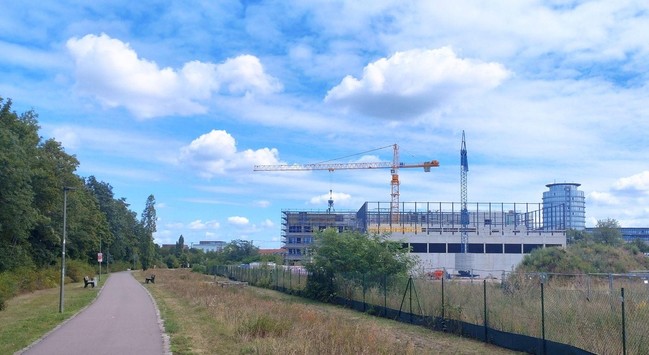 Bäume, Radweg und breiter Wiesenstreifen an einer Baustelle mit Kränen. Hohe Rohbaufassade mit großen, schwarzen Fensterluken nur im oberen Bereich. Blau-wolkiger Sommerhimmel und World-Trade-Center Dresden im Hintergrund. 