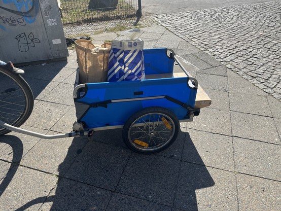 A blue grey bike trailer with a package sticking out the back and bags on the top 