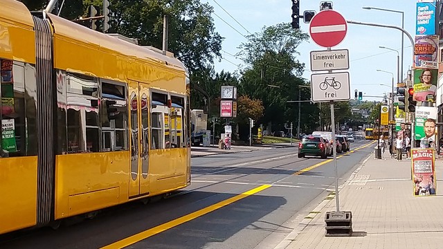 Links eine gelbe Straßenbahn kann nicht in die Haltestelle einfahren, weil vor ihr Autos stehen. Rechts am Fahrbahnrand ein rotes Einbahnstraße-Schild. Darunter der Hinweis 