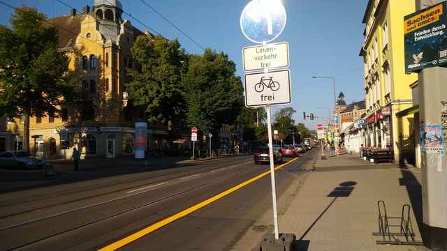 Breite Straße diagonal durch's Bild. Ein gelber Streifen trennt einen provisorischen Radweg ab. Autos fahren links davon. Ein Schild zeigt Einfahrt verboten, Linienverkehr frei, Fahrrad frei. Trotzdem fahren Autos selbstverständlich weiter.