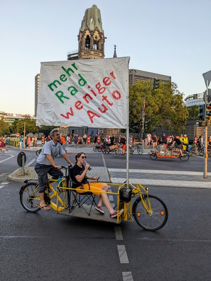 Im Vordergrund ist ein großes gelbes Lastenrad zu sehen, auf dem eine Person in entspannter Sitzposition Richtung Sonnenuntergang transportiert wird und dabei ein erfrischendes Kaltgetränk zu sich nimmt. Am Lastenrad ist eine Flagge mit der Aufschrift 