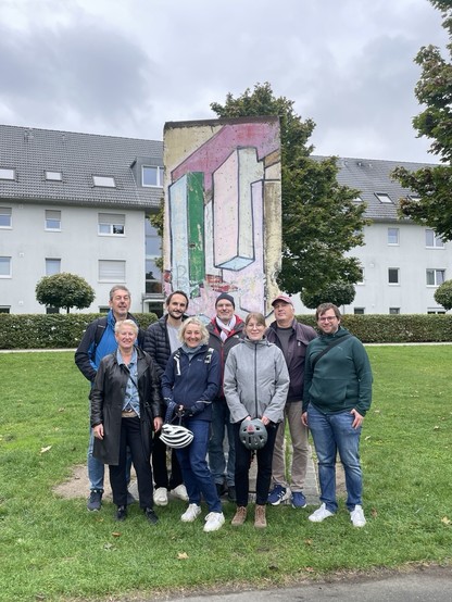 A group of nine people poses in front of a section of the Berlin Wall featuring colorful graffiti. They stand on a grassy area with residential buildings visible in the background under a cloudy sky. Some individuals are wearing jackets, and one holds a bike helmet.