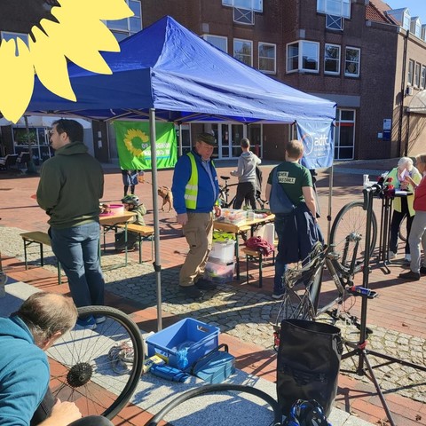 Ein blauer Pavillon mit Menschen von den Grünen und vom ADFC. Im Vordergrund arbeitet jemand an einem defekten Fahrrad und flickt den Schlauch des Hinterrades. Im Hintergrund sprechen Menschen miteinander.