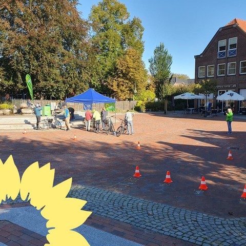 Ein Übersichtsbild über den Stand von Bündnis 90/Die Grünen und des ADFC auf dem Münzplatz in Stockelsdorf. Im Hintergrund ist ein blauer Pavillon zu sehen, an dem Menschen mit Fahrrädern stehen. Im Vordergrund markieren Verkehrshütchen einen Hindernisparcours.