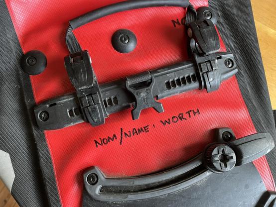 A red Ortlieb pannier with black clips. Mom/Name: Worth is written on it